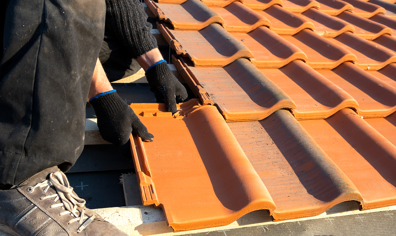 Closeup of worker hands installing yellow ceramic roofing tiles mounted on wooden boards covering residential building roof under construction. Learn how often how often to replace roof.